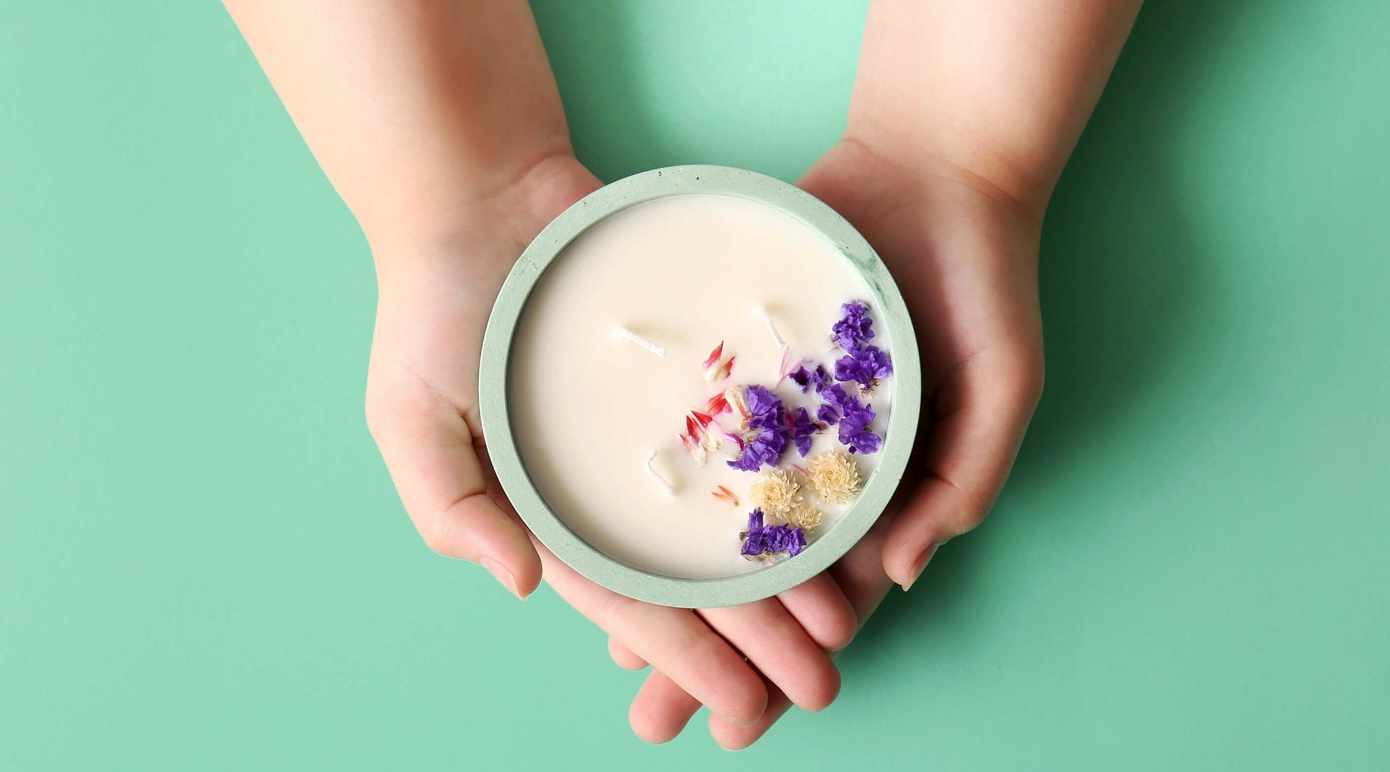 Hands holding a beautifully decorated soy wax candle with dried flowers on a mint green background.