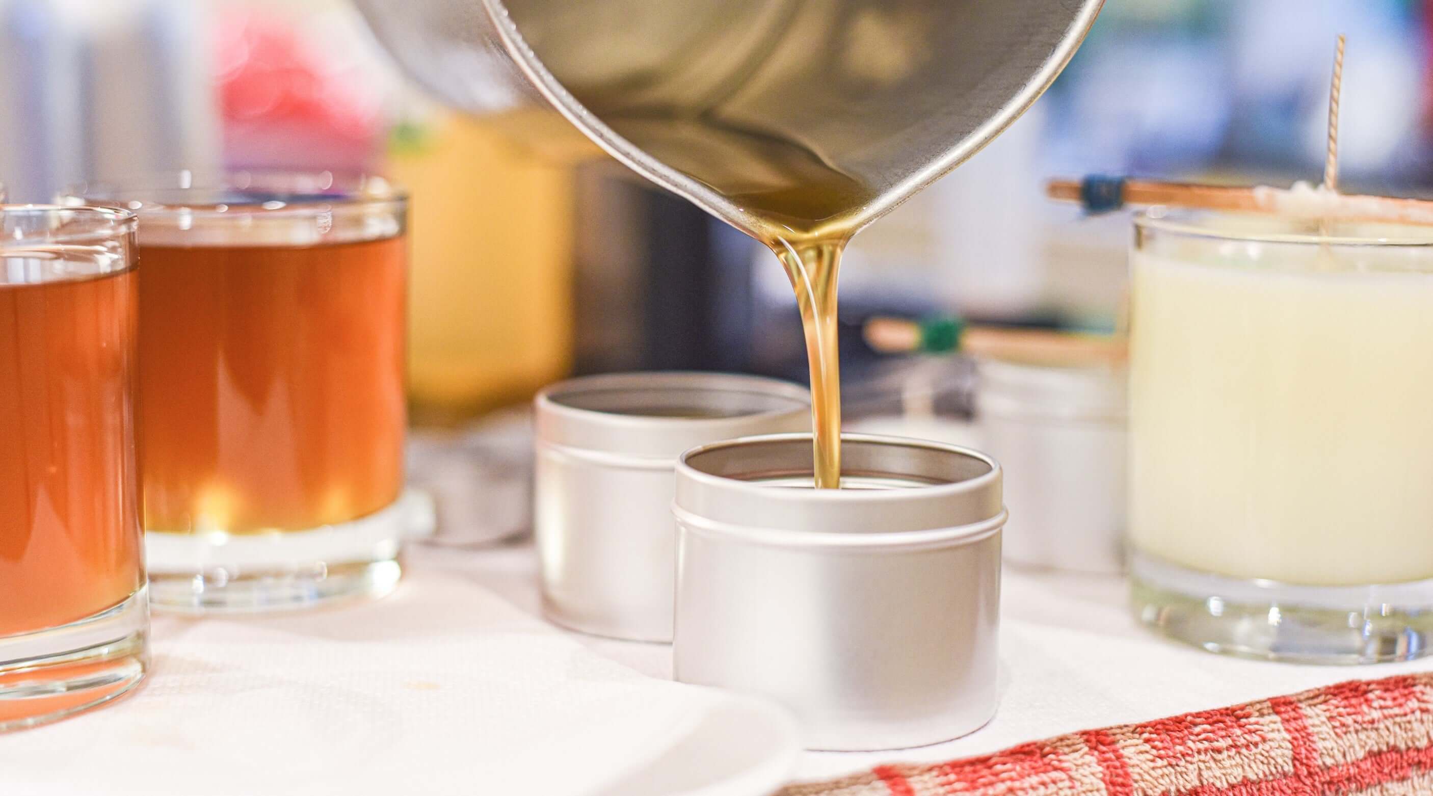 Pouring liquid soy wax into tin candle containers, surrounded by various candle jars on a table.