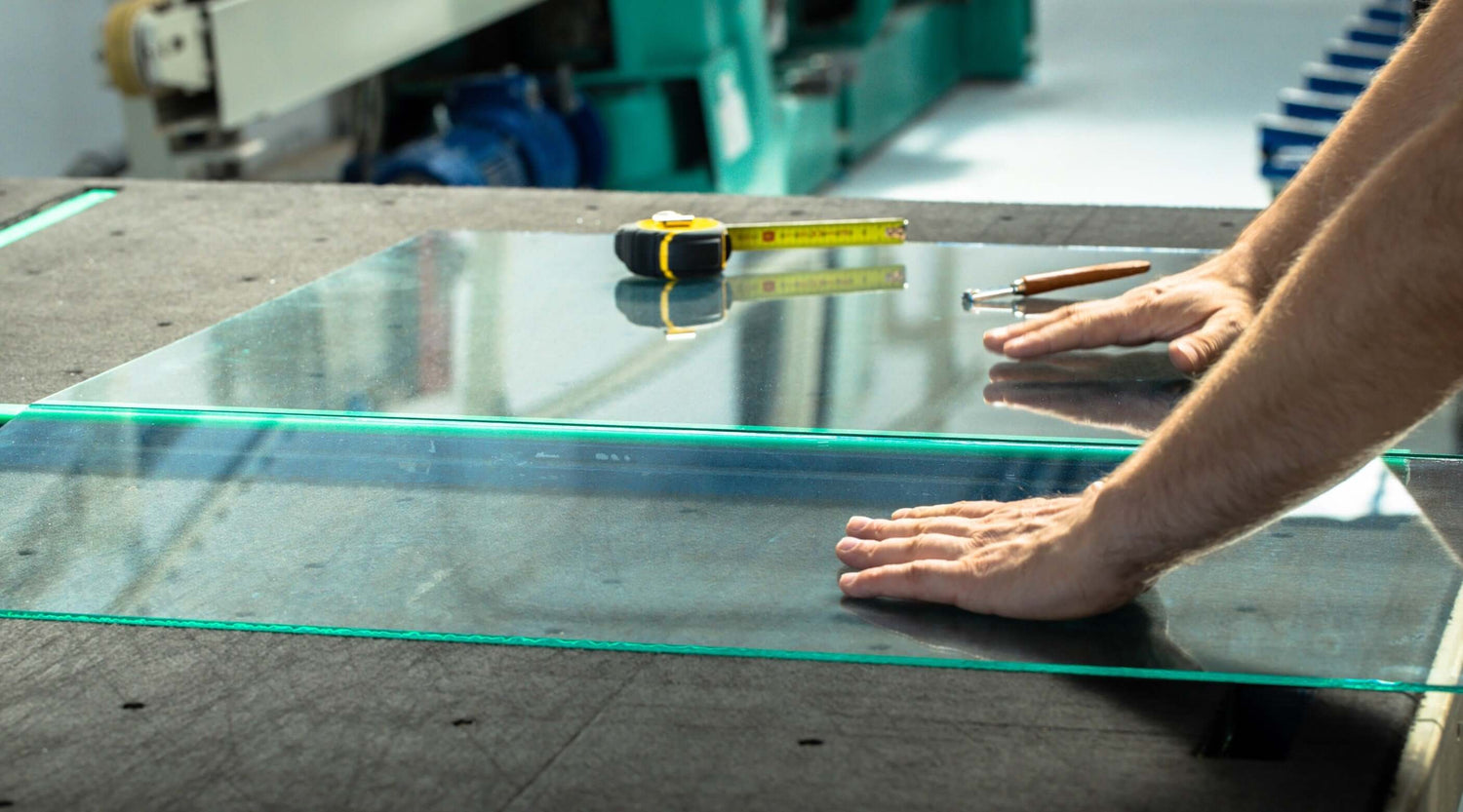 Person preparing to cut glass on a work surface with measuring tape and glass cutter for precise cutting.